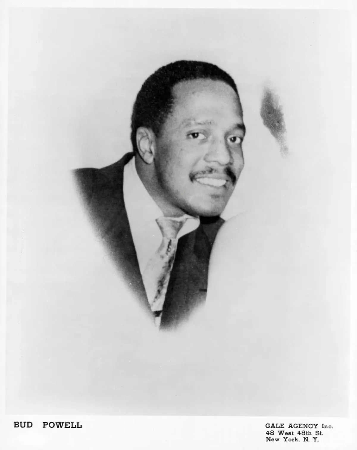 Black-and-white publicity portrait of Bud Powell in a dark suit and patterned tie, smiling; printed captions read “Bud Powell” and “Gale Agency Inc., 48 West 48th St., New York, N.Y.”