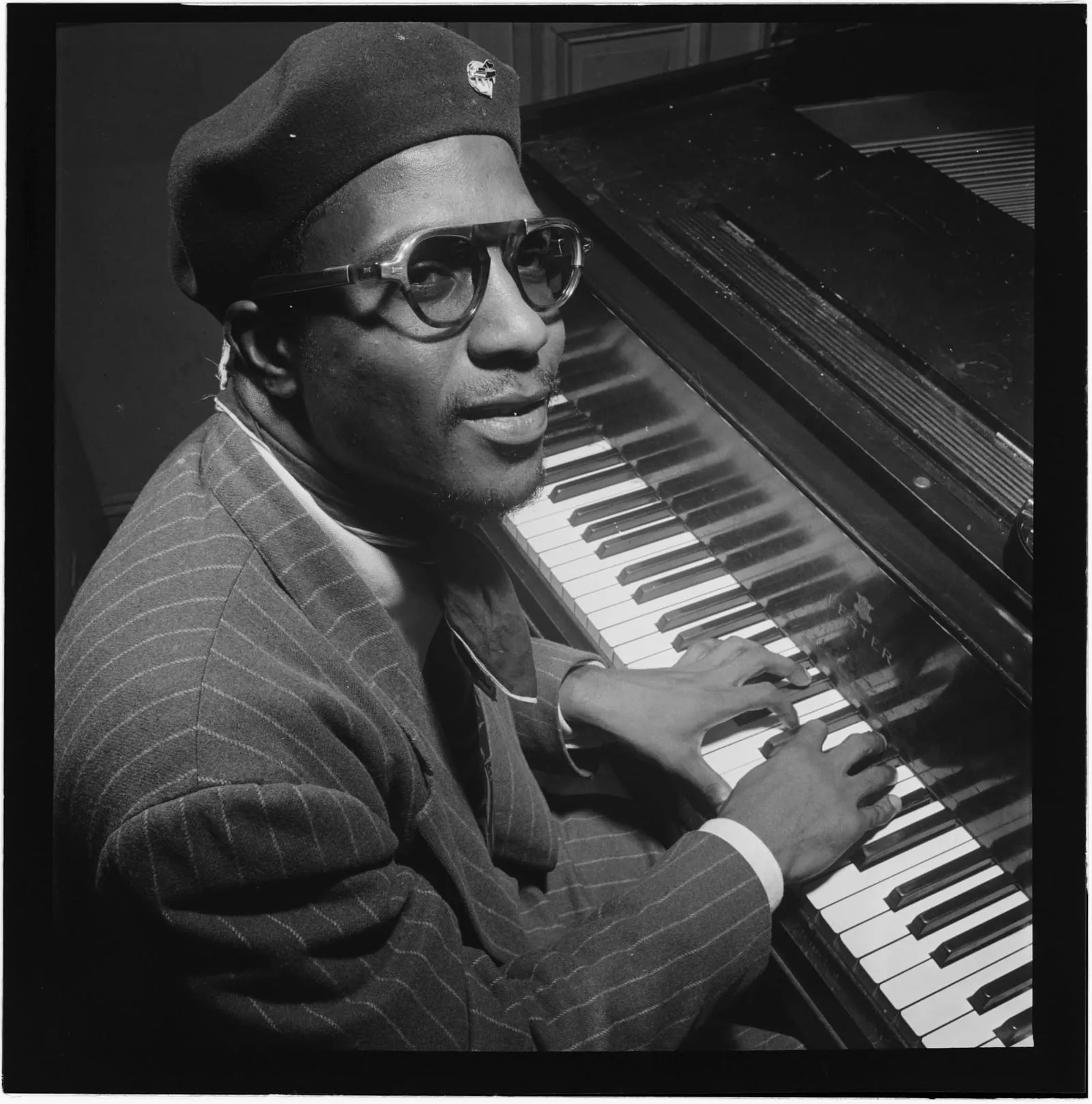 Thelonious Monk in a beret and horn-rimmed glasses, seated at a grand piano with his hands on the keys, looking toward the camera.