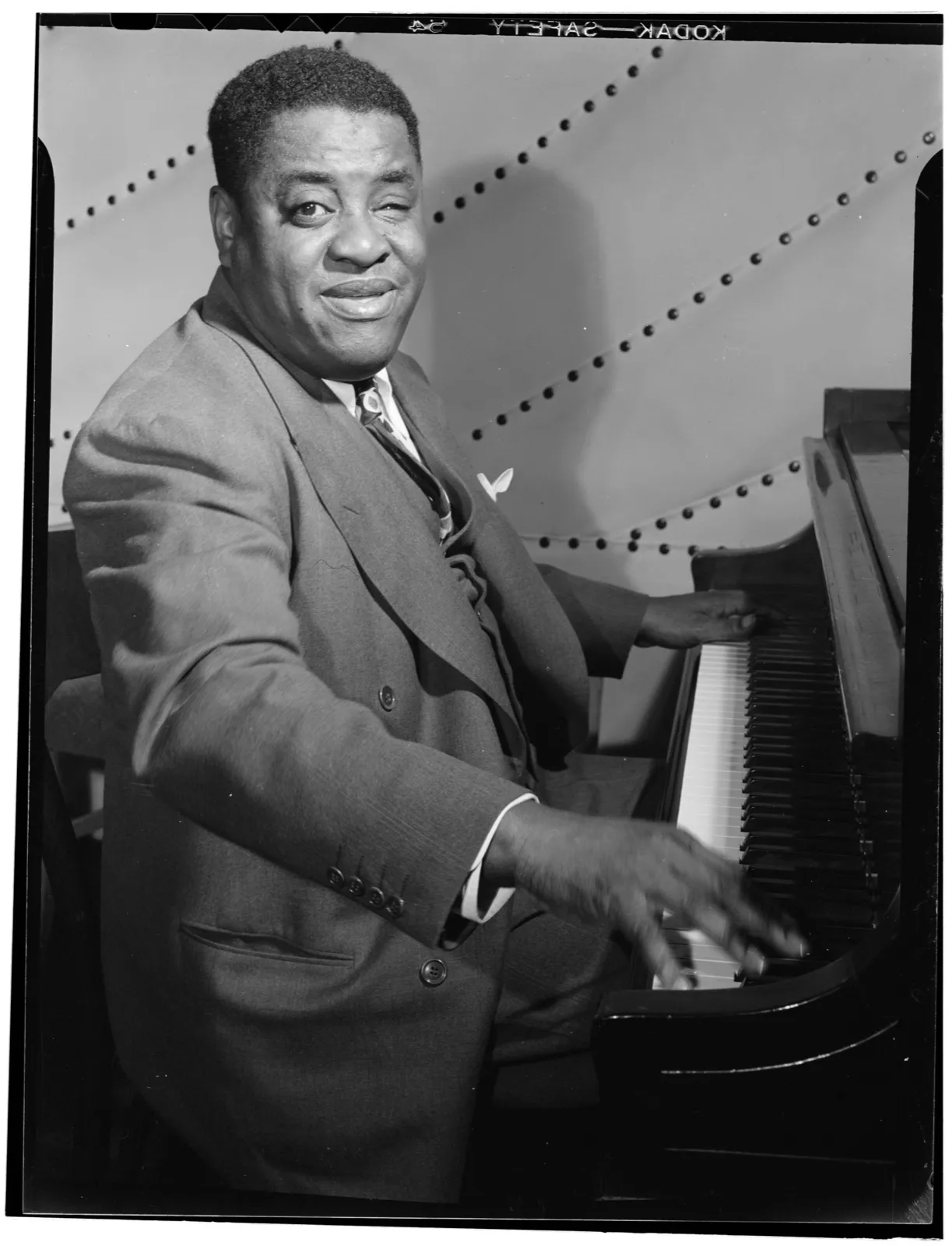 Art Tatum in a double-breasted suit, seated at a grand piano, turned toward the camera with a broad smile, his right hand on the keys.